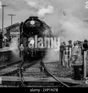 A vintage steam locomotive, 35028 Clan Line approaches the station  with smoke billowing from its chimney and a driver waving from the cab Stock Photo