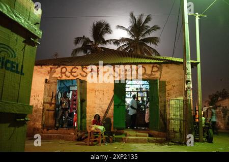 A small local shop in Gambia under moonlight with palm trees and street lights in the background, creating a serene and communal atmosphere. Stock Photo