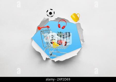 Ragged hole in paper with a shopping cart full of products emerging. Concept of shopping and buying various items through a creative perspectiv Stock Photo
