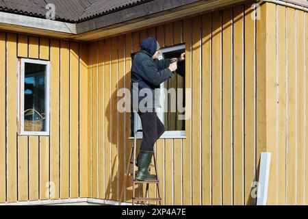 Roofing contractor does exterior trim of window opening while standing on steel step ladder, in front of facade of wooden country house on an autumn a Stock Photo