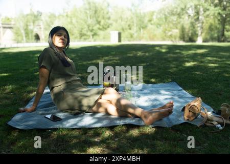 A woman is laying on a blanket in a park, with a bottle of beer and a cell phone nearby Stock Photo
