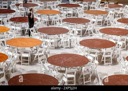 Rows of round dinner tables and chairs stand arranged in a bright hall waiting for guests, with a worker on the phone among them. Stock Photo
