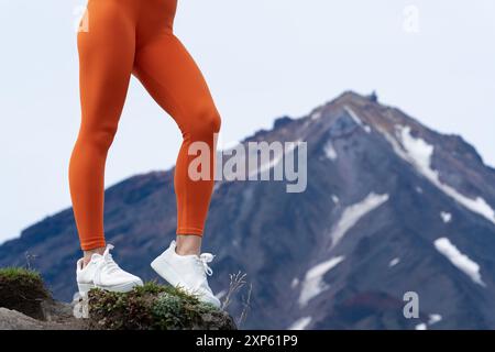 Woman dominates landscape with commanding presence as she performs challenging fitness routine in mountains, with volcanic peak in background. Close u Stock Photo