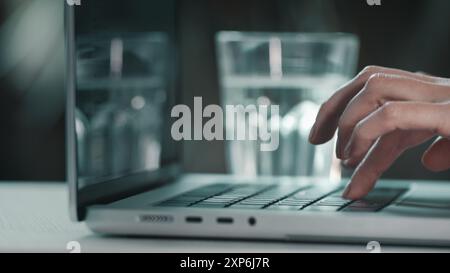 Female hands typing on computer close up. Woman user worker typing on laptop notebook keyboard desk working online with pc software apps. Drink water from glass, keep health balance hydration indoors Stock Photo