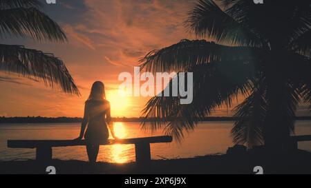 Young woman peacefully enjoys a stunning sunset over the ocean on a cozy beach bench. Bathed in warm golden hour sunlight. Creating a serene and idyllic atmosphere with swaying palm trees Stock Photo