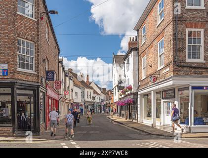 Historic buildings line a street and pedestrians walk between them. A pub is decorated with colourful hanging baskets. A sky with cloud is overhead. Stock Photo