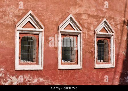 Three windows with metal cage in a row on the wall of an old red building. Ancient stone building. Simple decorative frame around the window. Stock Photo