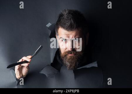 Barbershop advertising. Serious bearded man with straight razor looking through hole in paper. Bearded man, bearded male, vintage straight razor. Barb Stock Photo