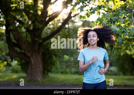 Young African American female athlete jogging outdoors in park. Woman running with sunlight filtering through leaves. Enjoying fitness and healthy lifestyle in natural environment Stock Photo