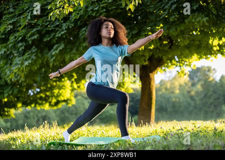 Young African American athlete doing yoga in warrior pose on yoga mat in park at sunset. Focused on stretching and balance in nature under tree with green leaves. Stock Photo