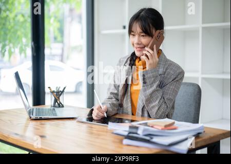 A busy young Asian businesswoman or female assistant is multitasking, talking on the phone with her client while working on her digital tablet at her Stock Photo