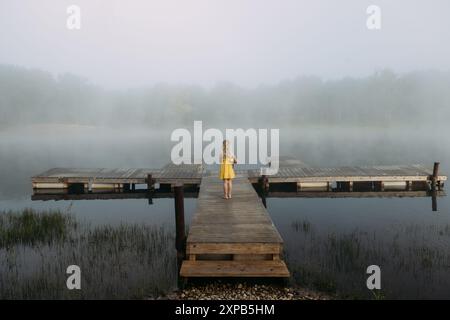 Girl in yellow dress stands on dock facing foggy lake in morning Stock Photo