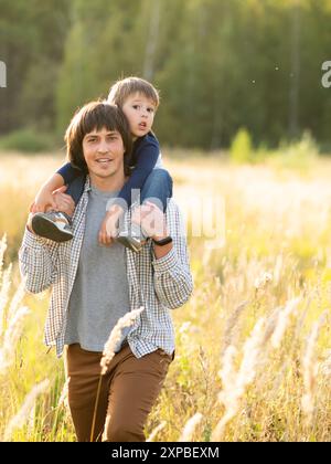 Father carries his son on shoulders. Family time outdoors. Man with child on autumn field at sunset. Dad and son explores nature together. Stock Photo