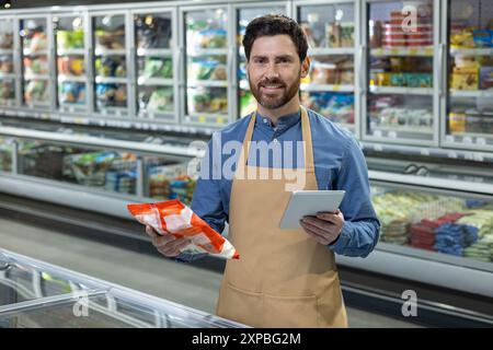Supermarket worker holding frozen food package and using digital tablet in front of refrigerated section. Smiling male employee in apron managing inventory. Modern grocery store environment. Stock Photo