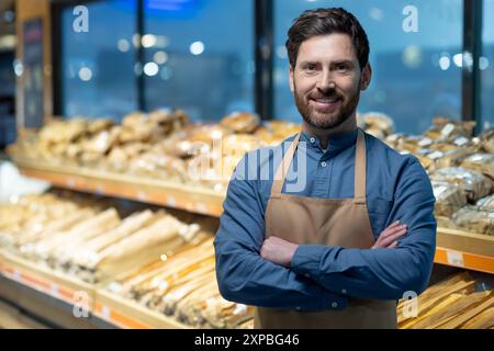 Confident baker standing with arms crossed in bakery shop surrounded by fresh bread. Person showcasing pride in craftsmanship and dedication to baking. Background displays variety of breads Stock Photo