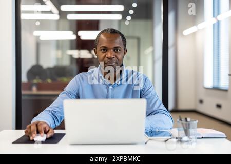 Confident businessman working on laptop in modern office. Focused professional using computer and mouse, performing work duties. Bright, modern office background. Stock Photo