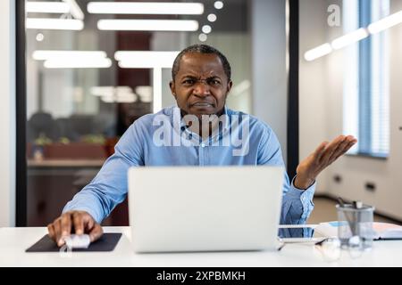 Businessman sitting at desk using laptop showing confused expression. Working professional in office environment, frustrated or perplexed. Technological or work-related issues. Stock Photo