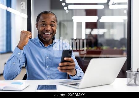 Businessman celebrating success using smartphone in modern office, showing excitement and achievement. Working at desk with laptop and other office supplies, portraying professional environment. Stock Photo