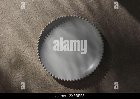 Summer restaurant still life scene. Blank white ceramic plate with decorative bobbin rim in sunlight on beige lime stone floor, table background Stock Photo