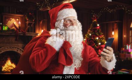 Happy Santa Claus with White Fluffy Beard and a Red Costume is Using a Smartphone. Accepting Online Gift Orders From All the Good Kids Around the World. Old Santa Holding a Bag with Presents Stock Photo