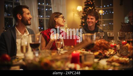 Handsome Boyfriend Presenting a Festive Gift to His Beautiful Surprised Girlfriend. Family and Friends Gather at Home in the Evening for a Traditional Christmas Dinner with a Turkey Roast Feast Stock Photo