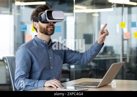 Businessman using virtual reality headset working on laptop in office. Exploring VR technology, innovative interaction, workplace creativity. Casual wear reflecting modern work environment Stock Photo
