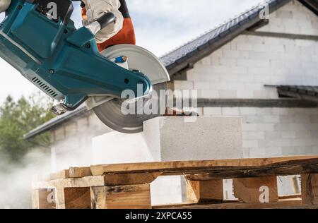 A worker uses a power tool to cut concrete blocks on wooden pallets at a construction site under a clear sky. Stock Photo
