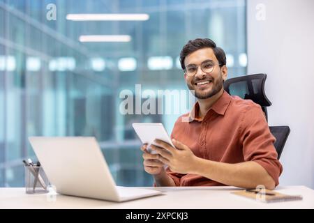 Smiling young professional man sitting at desk using tablet and laptop in modern office environment. Workspace includes notebook and phone. Concept of technology, business, productivity Stock Photo