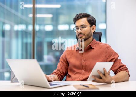 Serious businessman working with laptop and tablet in modern office. Focused on tasks, using both devices to enhance productivity. Professional setting with large windows and natural light. Stock Photo