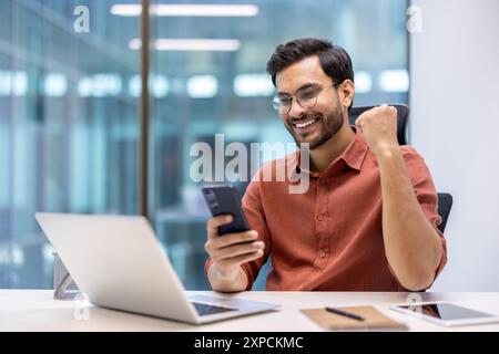 Excited businessman celebrating success at work using smartphone. Young professional sitting at office desk with laptop, phone, and notebook, showing joyful expression and happiness. Stock Photo