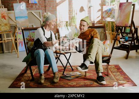 Senior Caucasian female artist sitting on chair in studio showing drawing in sketchbook to young man Stock Photo