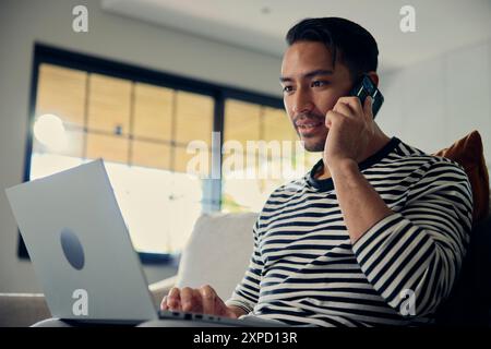 Man on couch using laptop, talking on cell phone, gesturing with arm Stock Photo