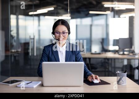 Asian businesswoman wearing a suit and headset while working at her office desk with a laptop. She is smiling and appears focused. Bright and modern office environment. Stock Photo