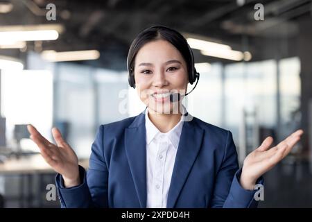 Confident businesswoman wearing a headset, looking at the camera, and talking on a video call. She is gesturing with her hands in a modern office environment. Stock Photo