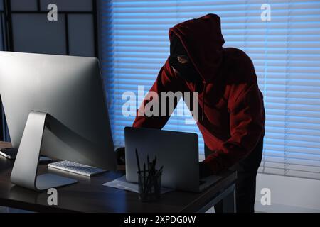 Thief wearing balaclava stealing laptop in office at night. Burglary Stock Photo
