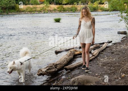 A woman in a white dress walks her white Samoyed husky dog on the shores of the Willamette River Stock Photo