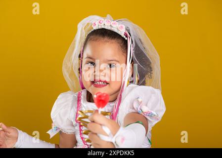 Portrait of a beautiful and cheerful girl in a wedding dress, showing a red lollipop to the camera. Isolated on a yellow background. Stock Photo