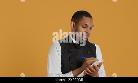 Professional caterer writing clients orders on notepad for the bill, takes notes of desired food against yellow background. Waiter with apron, notebook service for customer satisfaction. Camera A. Stock Photo