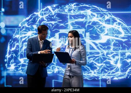 Team of engineers analyzing AI models with large digital brain displayed behind. IT staff members, using machine learning to better understand and develop artificial intelligence Stock Photo
