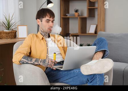 Young handsome man smoking disposable electronic cigarette and using laptop on armchair at home Stock Photo