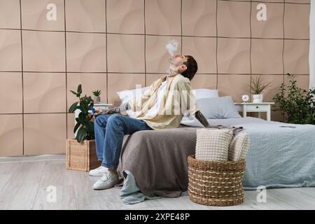 Young man smoking disposable electronic cigarette and resting on bed at home Stock Photo