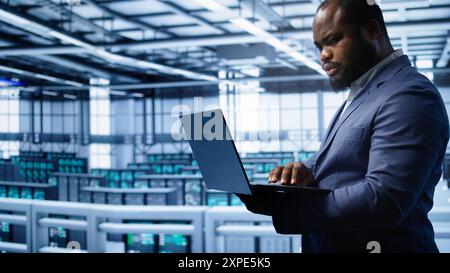 Software developer in server hub checking recovery plan on laptop, monitoring hardware energy consumption. African american worker uses notebook in data center, ensuring rigs are functioning, camera B Stock Photo