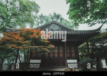 A traditional Chinese garden pavilion with a tiled roof surrounded by lush green trees, some with red foliage. Stock Photo