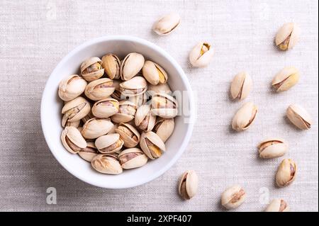 Pistachios in their shells, roasted and salted, in white bowl on linen. Crunchy roasted fruits of Pistacia vera, with opened shells and seeds within. Stock Photo