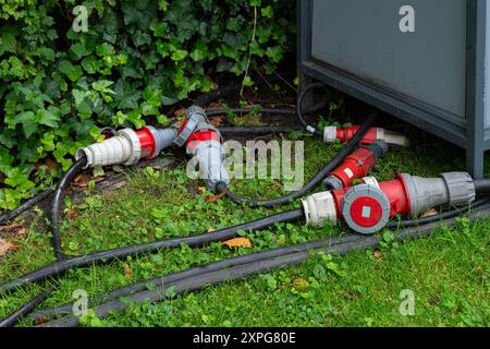 Heavy-duty electrical connectors and cables lie on the grass near a construction panel, surrounded by greenery. Used for powering construction equipment. Stock Photo