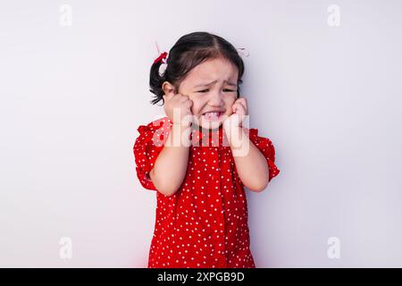 Asian toddler girl with both hands on cheeks, wearing a red polka dot dress and red and white ribbons on hair isolated on white background. Stock Photo