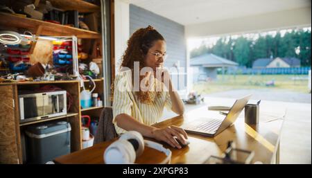 Happy Hispanic Female Engineering Student Sitting At Desk And Using Laptop Computer In Opened Garage. Woman Studying, Doing Research For New Project, Reading Articles, Watching Online Courses. Stock Photo
