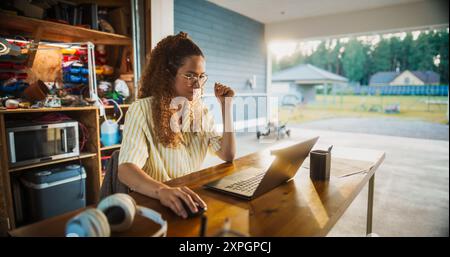 Young Hispanic Female Software Engineer Programming On Laptop Computer In Opened Garage. Woman Running A Startup With Innovative SaaS Solution For Businesses. Developer Writing Code, Optimizing System Stock Photo