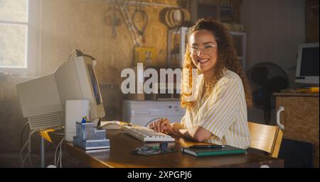 Hispanic Female Software Engineer Programming On Old Desktop Computer In Retro Garage, Looking At Camera And Smiling. Woman Starting an Innovative Startup Company In Nineties. Nostalgia Concept. Stock Photo