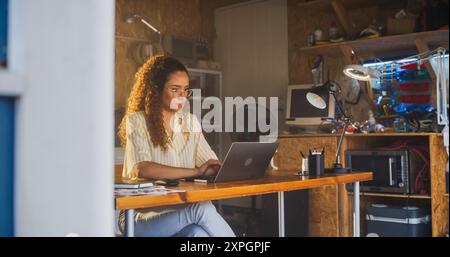 Hispanic Female Programmer Writing Code On Laptop Computer In Garage At Home. Woman Working On Innovative Software As A Service Startup For Business. Digital Entrepreneur Starting Company Concept. Stock Photo